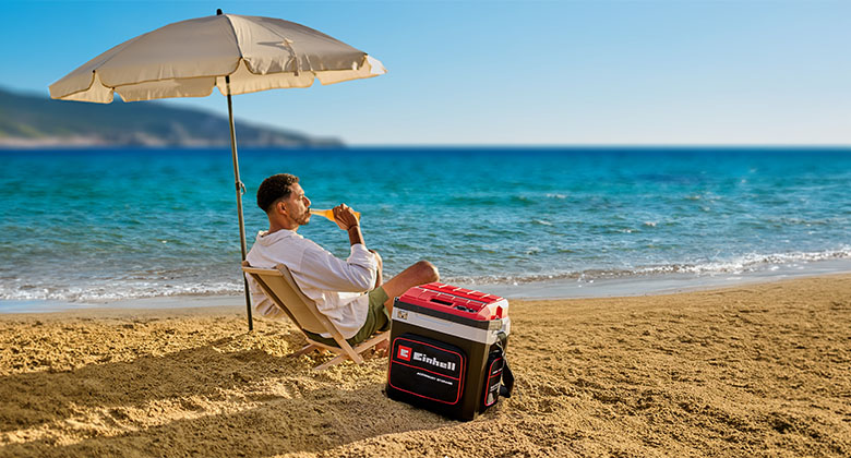 Mann sitzt am Strand unter Sonnenschirm neben einer Einhell Kühlbox mit rotem Deckel vor dem Meer.