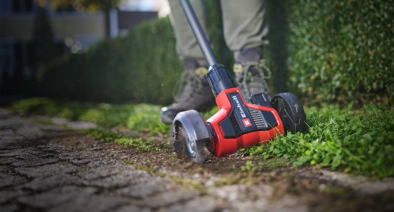Der Einhell Akku-Fugenreiniger entfernt Unkraut aus Pflasterfugen entlang eines Gartenwegs.
