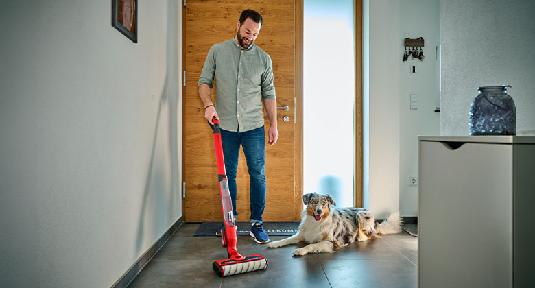 Mann reinigt den Flur mit rotem Akku-Hartbodenreiniger, neben ihm liegt ein Hund auf dem Boden.